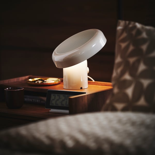 Close-up of sa bedside table with BLOMPRAKT bluetooth speaker lamp, being the only light source in the otherwise dark room.