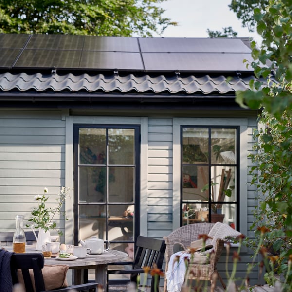 A patio in front of a small house with light gray wood paneling and two glass doors with black frames. On the roof are solar panels above dark gray roof tiles. In the foreground is a round table with chairs, set with glasses, a carafe, and a vase with green twigs.