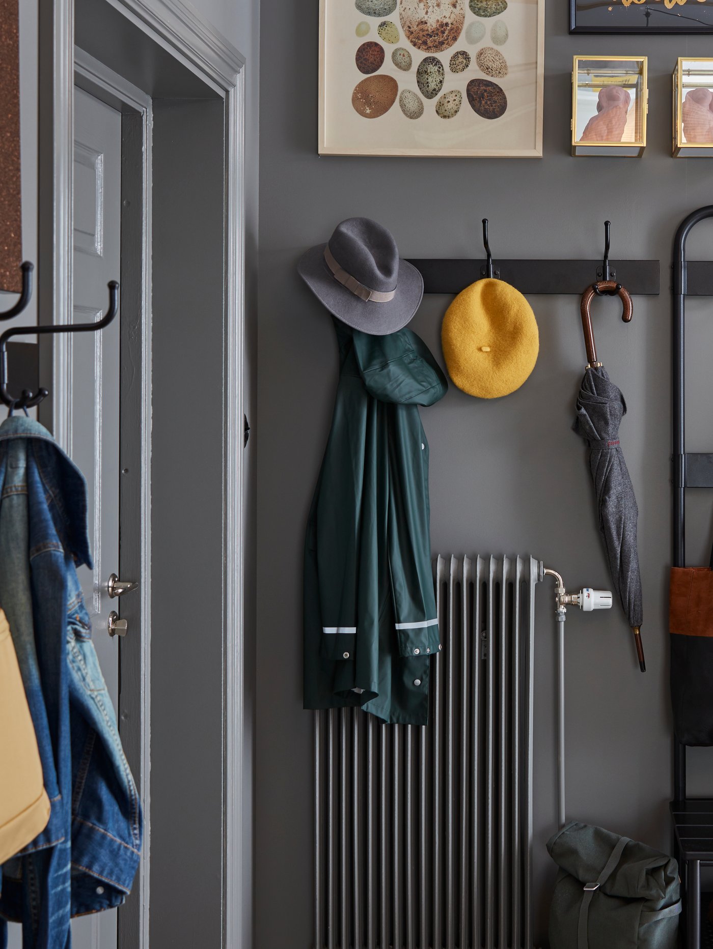A PINNIG rack with 3 hooks on a grey wall by a door placed above a radiator and holding jackets, hats and an umbrella.