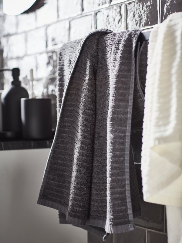 Draps de bain VÅGSJÖN blanc et gris foncé sur un porte-serviettes noir sur un mur en carrelage gris, plus un ensemble d’accessoires 3 pièces en grès.