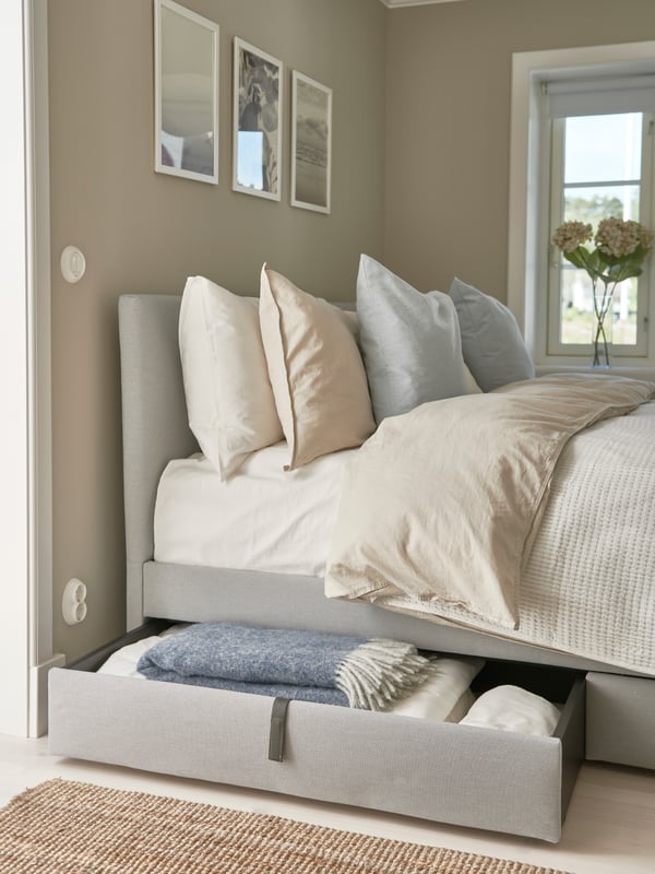 Sovrum med bäddad säng i vitt och ljusgrått, stoppad sängram med utdragen förvaringslåda fylld med filtar, tre tavlor ovanför sängen, beige väggar, fönster med ljusinsläpp och vas med blommor på fönsterbrädan.