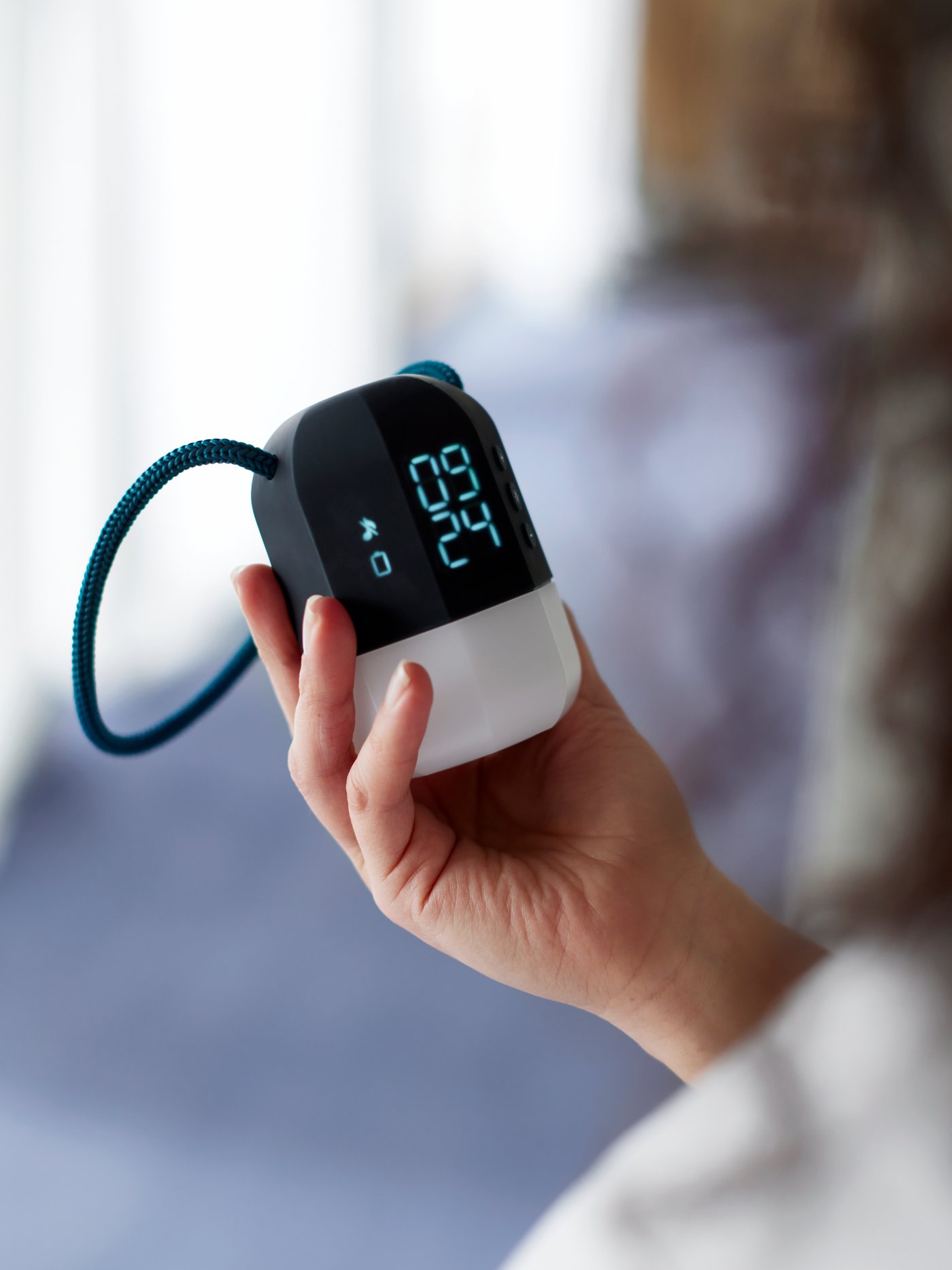 A hand holding a FNURRA digital alarm clock and wake-up light in a bright bedroom with white curtains by the windows.