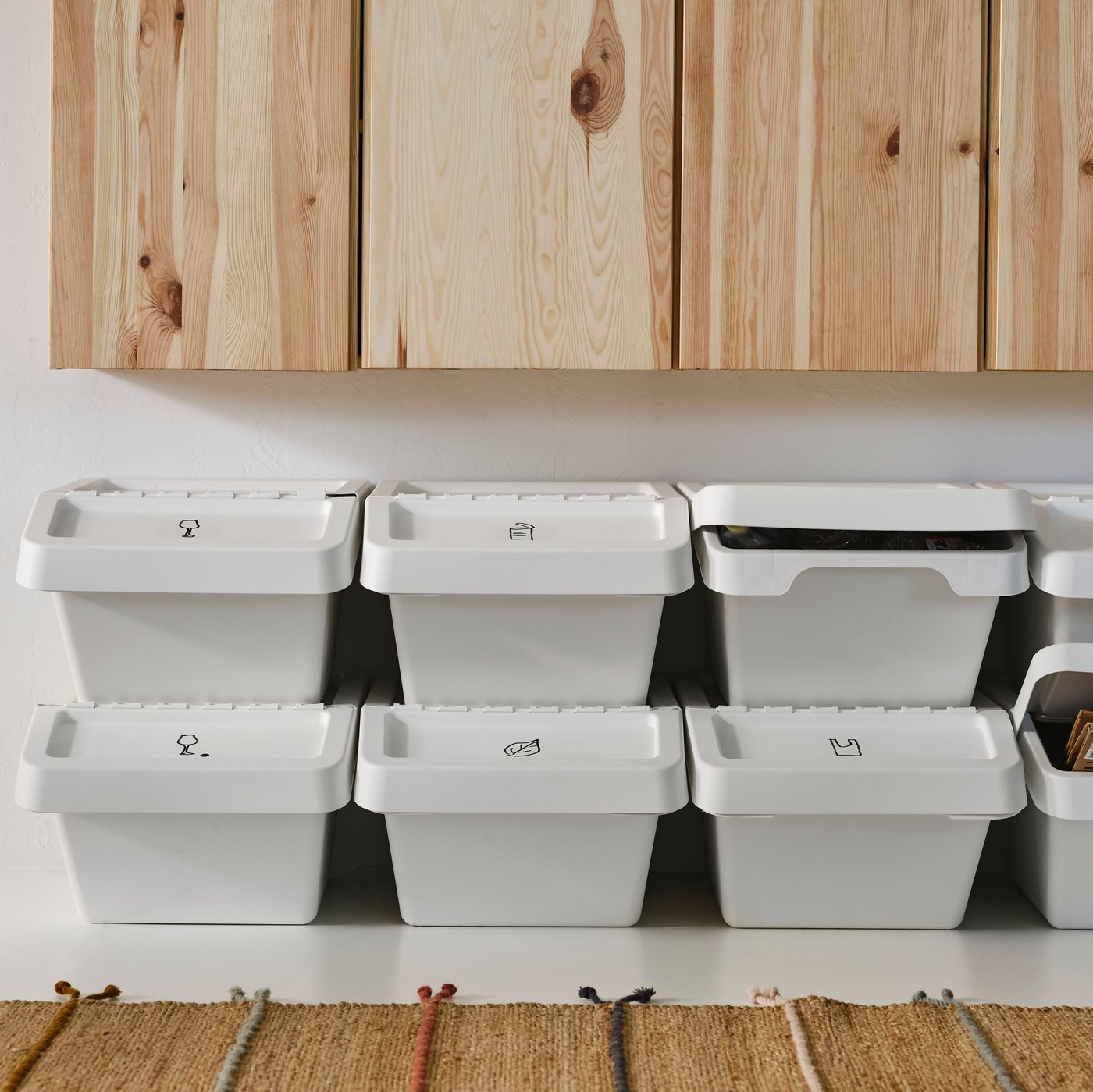 Rows of white SORTERA waste sorting bins with lids stacked on top of one another under a row of pine cabinets, plus a rug.