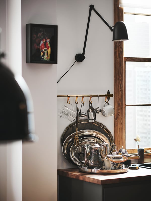 A wooden worktop with a tray of antique silverware on top. Above, there is a kitchen rail with utensils hanging from it.