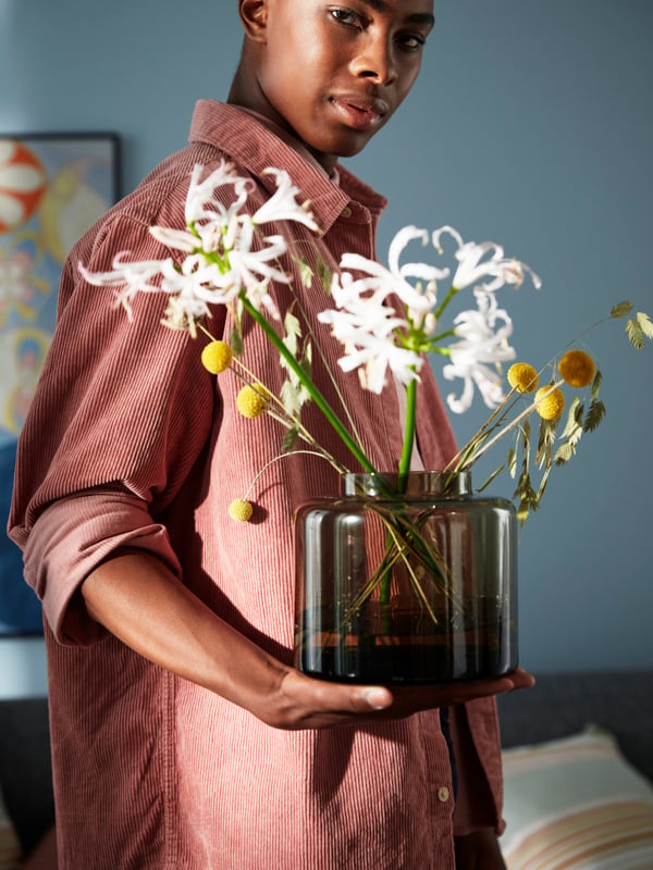 A man holding a brown KONSTFULL vase in glass with some white and yellow flowers against the backdrop of a living room.