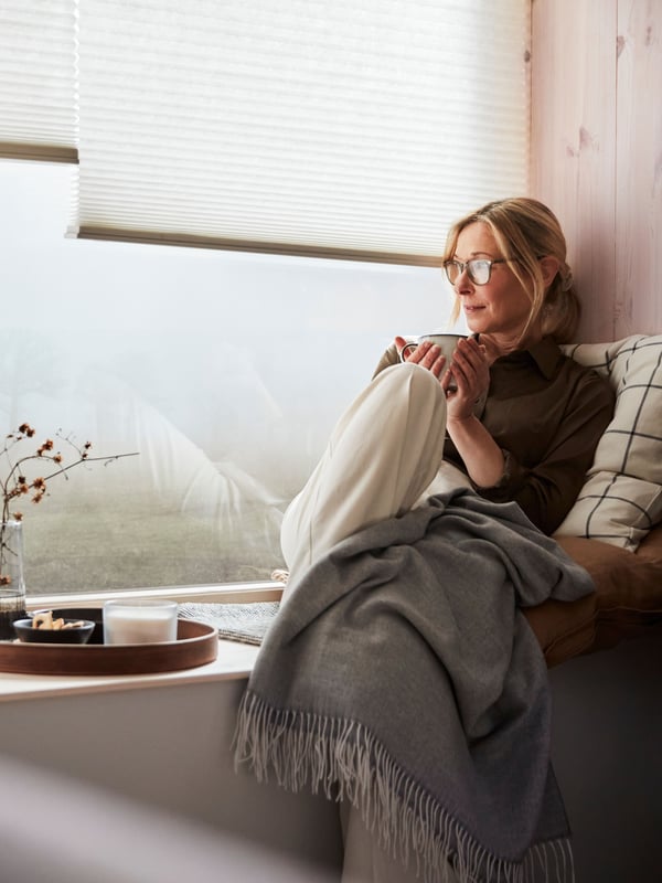 A person lounges on cushions on a windowsill. Two white PRAKTLYSING cellular blinds hang halfway down in the window.