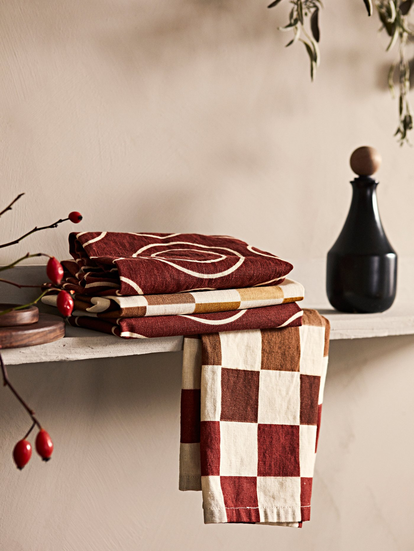 Folded KRÖSAMOS tea towels together with a KRÖSAMOS black bottle on a wooden shelf inside a kitchen.