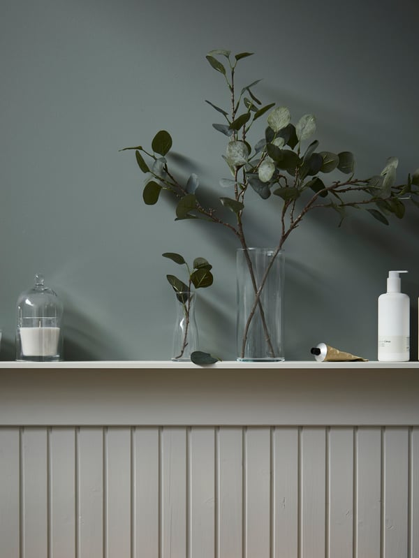 A half-panel wall with a shelf with a candle in a glass dome, and artificial olive twigs are displayed in glass vases.