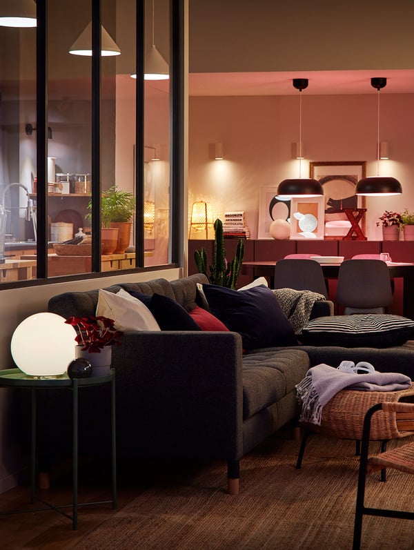 A room with a sofa, a footstool, a table and chairs, with a lamp on a side table and hanging lighting in the background.