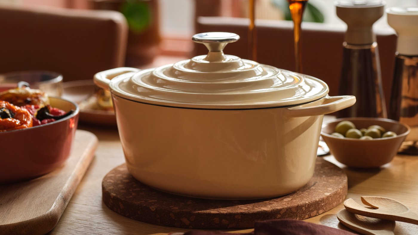 A VARDAGEN pot in enamelled cast iron in light yellow stands on a wooden dining table surrounded by food and candles.