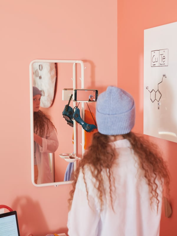 A girl in a beanie standing in front of a white MÖJLIGHET mirror. A phone in camera mode rests in the mirror’s phone holder.