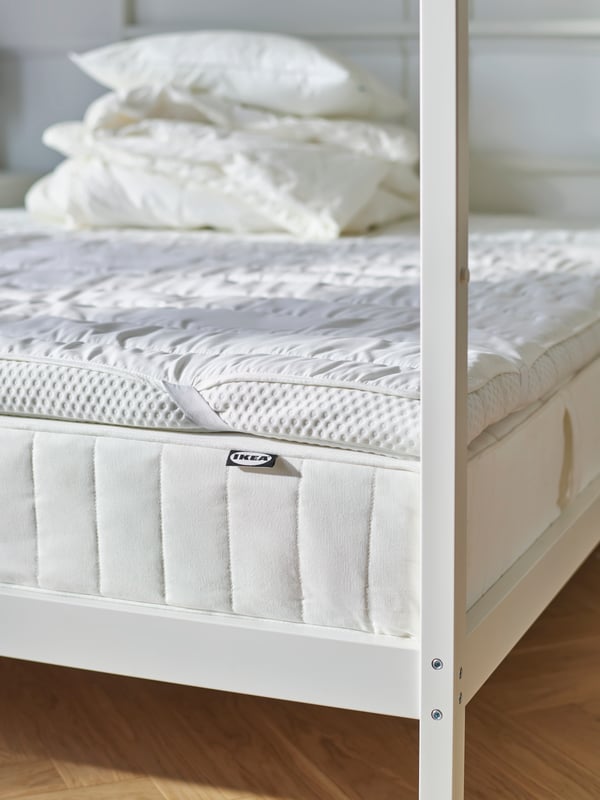 Lit à colonnes VITARNA et ses différentes couches côté matelas. Une pile de couettes et d’oreillers est posée sur ce dernier.