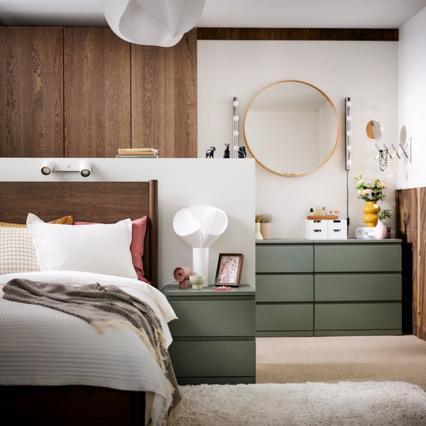 Deux commodes STORKLINTA gris vert avec 3 tiroirs dans un coin d’une chambre équipée d’un lit et d’une table de chevet devant.