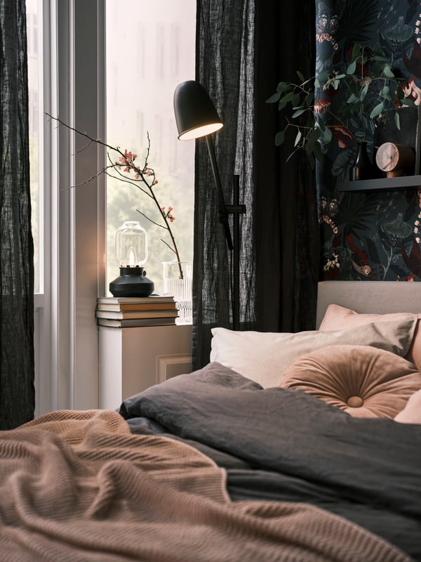 A black SKURUP floor/reading lamp stands beside a bed with a dark grey DYTÅG duvet cover and a pale pink INGABRITTA throw.