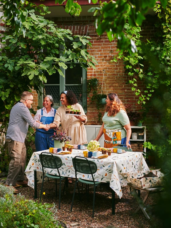 A garden with four people around a table with plants, DAKSJUS trays, plant pots and accessories on a DAKSJUS tablecloth.