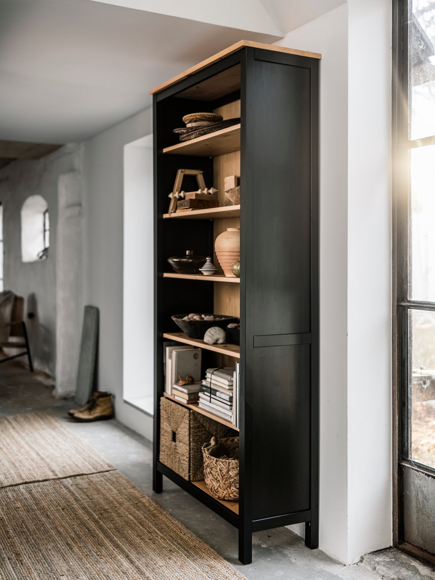 Et lyst rum med en sort/brun/lysebrun HEMNES bogreol med pyntegenstande, bøger og kurve på hylderne.