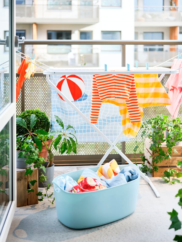 A blue TORKIS laundry basket filled with laundry and a MULIG drying rack on a sunny balcony decorated with plants.
