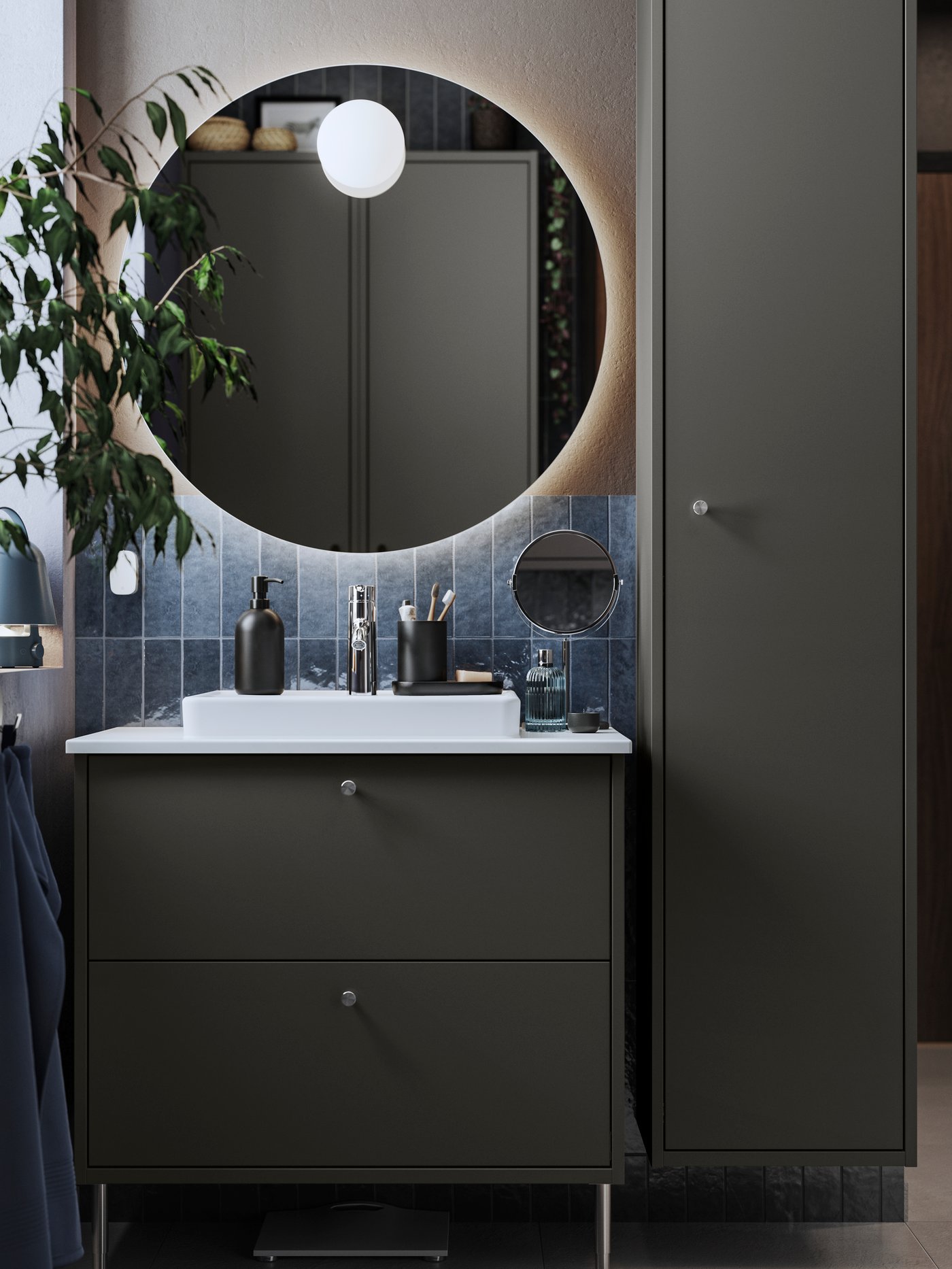 A dark grey HAVBÄCK washstand and a round FAXÄLVEN mirror with built-in lighting are placed in a dark-tiled bathroom.