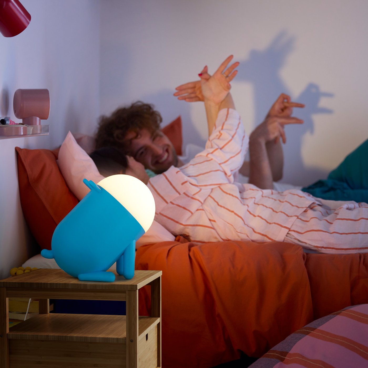 Two people lie in a bed and make shadow puppets in the light cast by a dog-shaped GREJSIMOJS LED table lamp in blue.