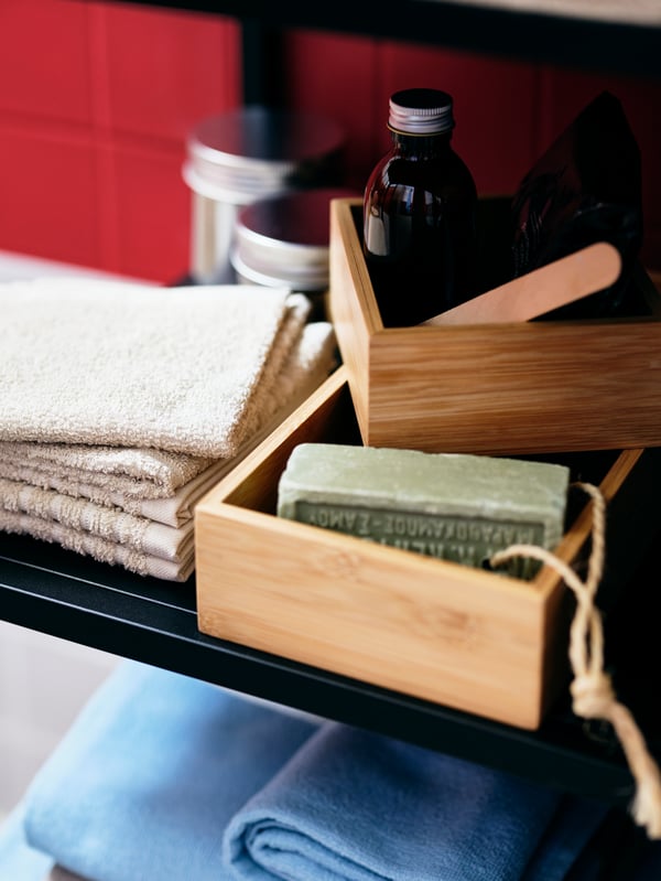 Folded towels, jars and bottles, and bathroom accessories in a pair of TAVELÅN trays share a shelf of an ENHET cabinet.