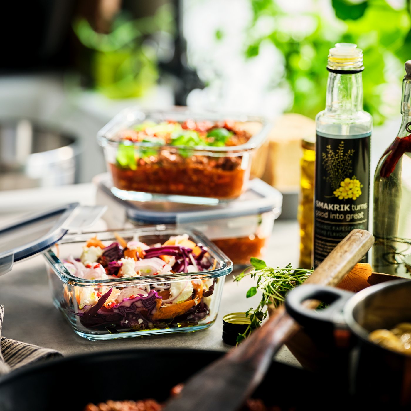 Glazen IKEA bewaarbakjes met kleurrijke salades staan op een zonnig keukenblad, omringd door verse kruiden, een fles olie en een pan.