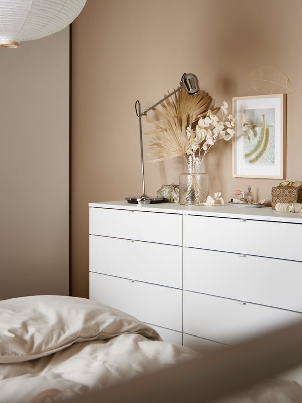 Minimalistisches Schlafzimmer mit einer weißen VIHALS Kommode, Dekoartikeln, Trockenblumen in einer KONSTFULL Vase, einer MODERMOLN Schreibtischlampe und weicher Bettwäsche. Der Raum strahlt eine ruhige, gemütliche Atmosphäre aus.