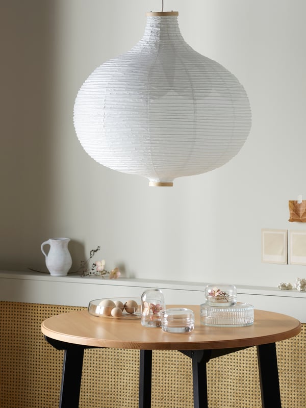 A large RISBYN paper pendant light hangs above a circular wooden coffee table with clear glass vases and dishes. 