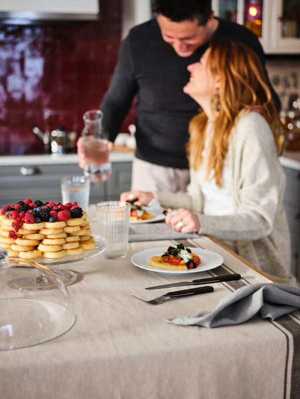 Due persone accanto a un’isola per cucina con tovaglia e bicchieri VARDAGEN, cibo sui piatti e alzata ARV BRÖLLOP in vetro.