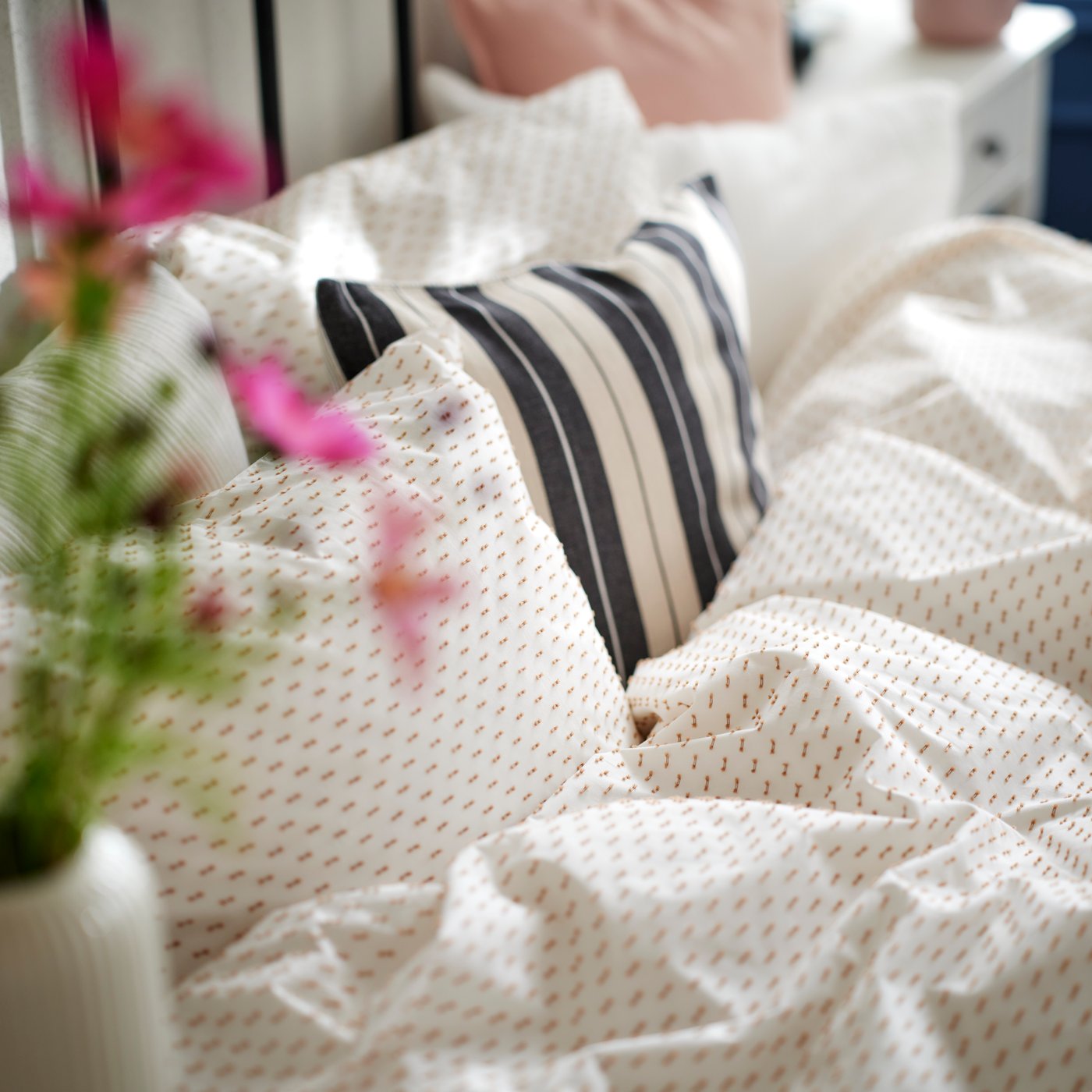 Ein Bett ist mit einer weiß-beigen BANDKNIPPE Bettwäsche bezogen. Passend dazu liegt ein dekoratives Kissen mit gestreiftem KORALLBUSKE Bezug im Bett.