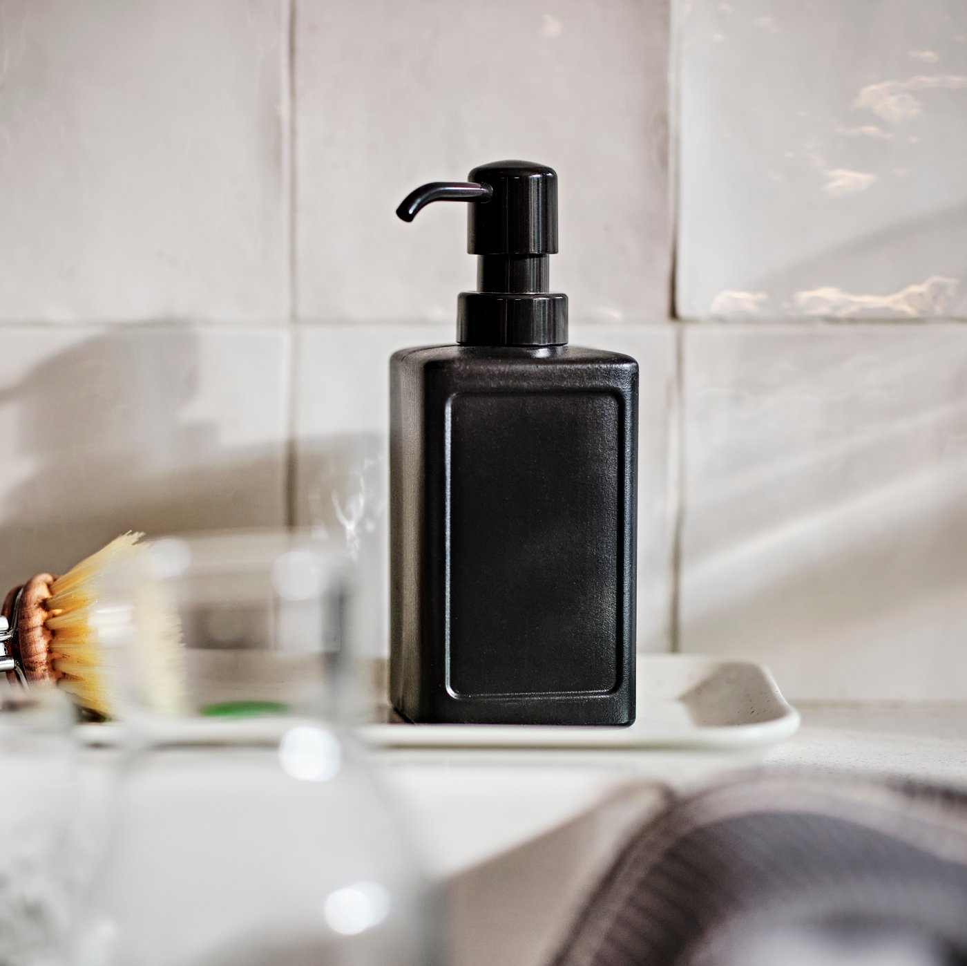 A grey RINNIG soap dispenser on a small white tray on a worktop in a kitchen against a white tiled wall.