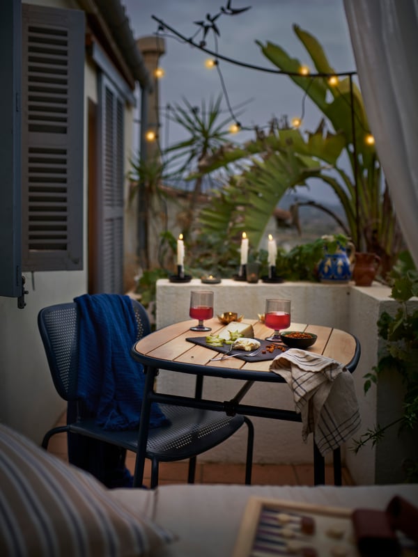 A DUVSKÄR chair stands beside a DUVSKÄR table laid with a cheese platter and two glasses in a lush outdoor space at dusk.