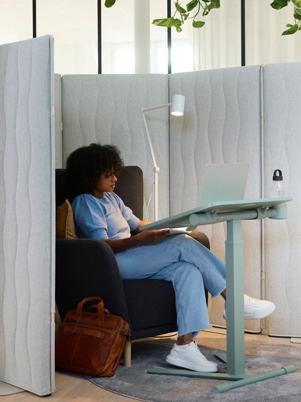 Une personne est assise dans un fauteuil gris foncé devant une table pour ordinateur portable à roulettes MITTZON verte inclinée vers le bas sur laquelle est posé un ordinateur portable.