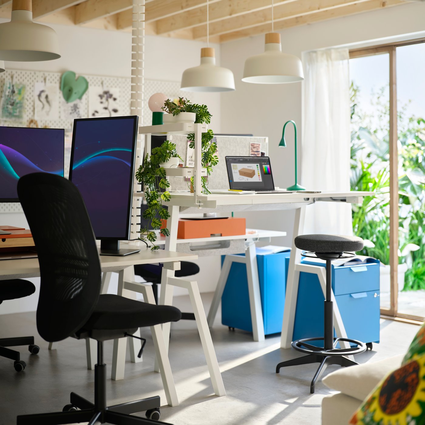 Bright start-up office with white sit/stand desks, large monitors, ergonomic seating, and blue storage cabinets on casters.