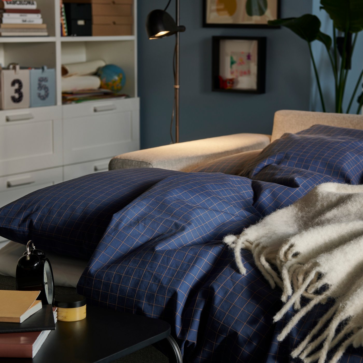 A softly lit living room with a BÅRSLÖV sofa-bed made with DOFTAKLEJA bed linen and an OLSERÖD side table by the bed.