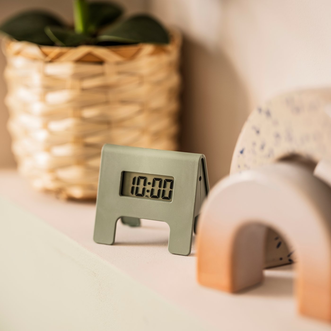 A softly sunlit space with a green KUPONG alarm clock nestled between decorations and a leafy plant in a woven plant pot.