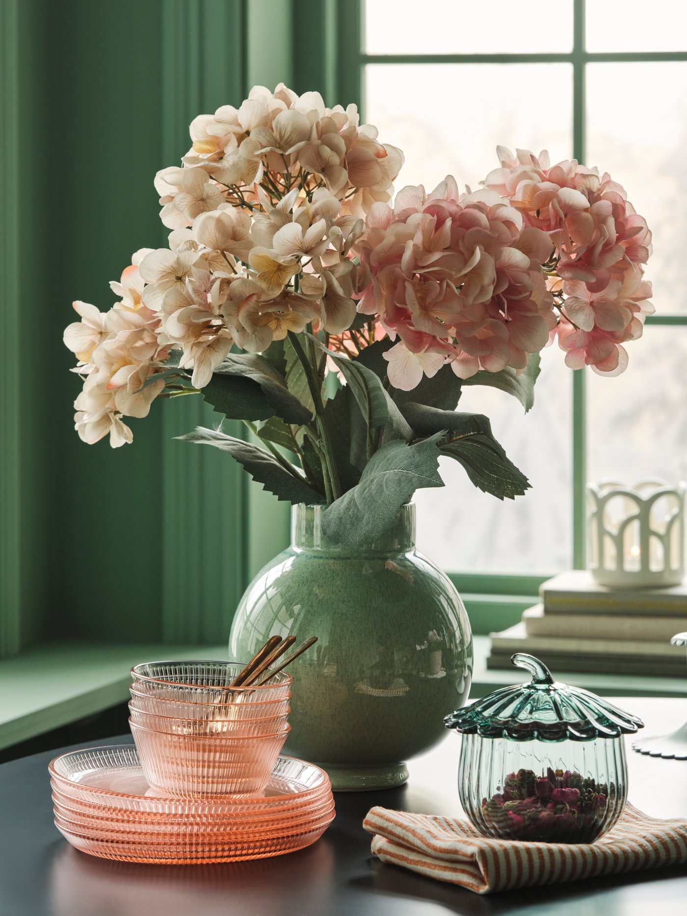 A bouquet of SMYCKA artificial flower in light pink and beige stand in a green vase on a table with glass plates and bowls.