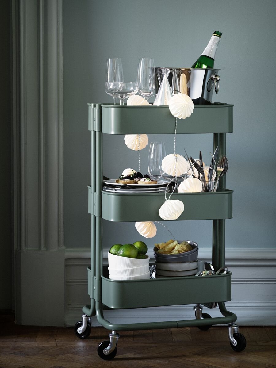 Un chariot vert RÅSKOG est rempli d'articles pour une fête, comme des verres, de la vaisselle et des boissons à partager.
