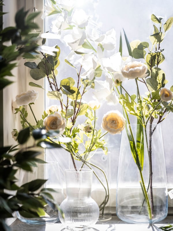 Quatre vases en verre de formes différentes contenant des fleurs blanches artificielles et des branches vertes feuillues, sur le bord d’une fenêtre.