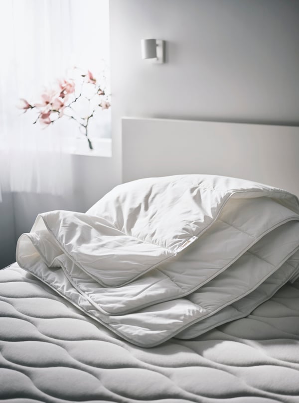 A SMÅSPORRE light warm duvet folded up on an unmade mattress on a bed in a white room by a window with a vase on the sill.