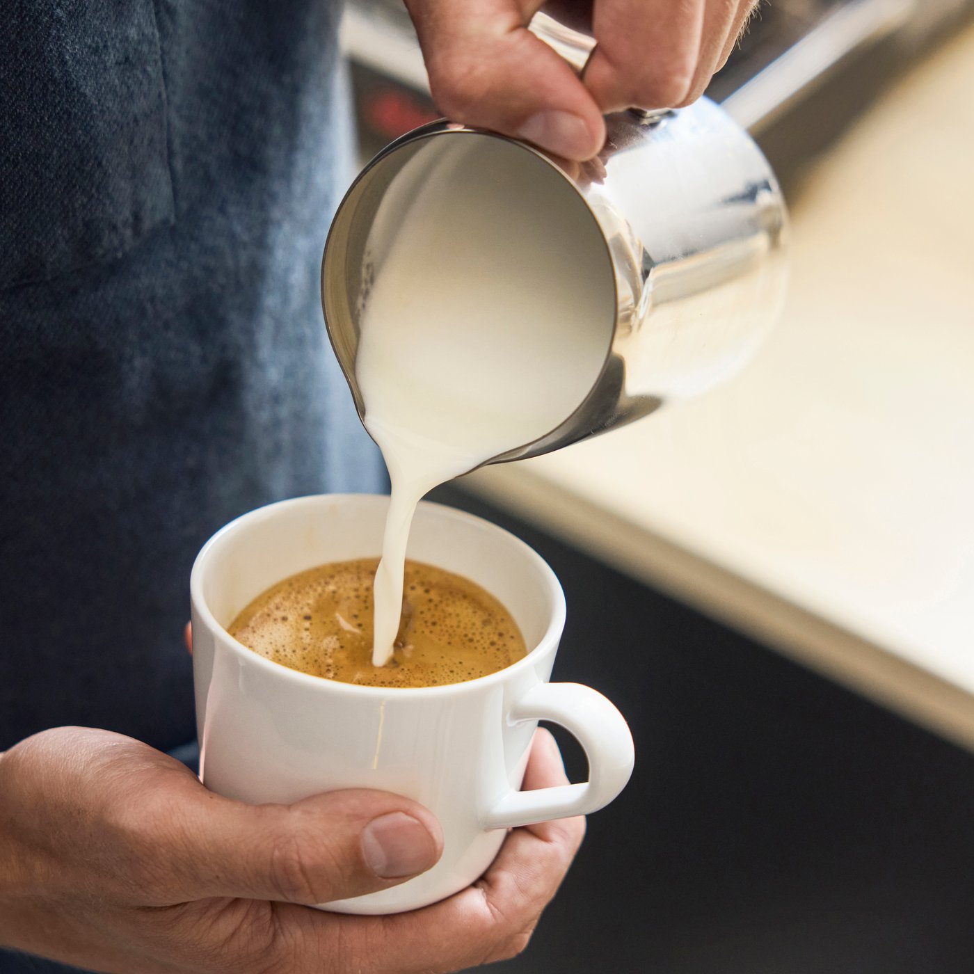Milk poured from small pitcher into coffee cup, creating foam, with hands holding cup and pitcher in café setting.