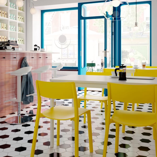 An airy coffee-bar interior with white JANINGE bar stools by a counter and yellow JANINGE chairs at white TOMMARYD tables.