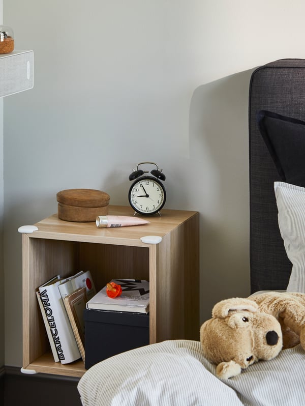 Un módulo EKET de color madera utilizado como velador, con las esquinas protegidas por protectores UNDVIKA. A un costado, sobre la cama se ve un peluche de perro GOSIG GOLDEN sobre una funda de plumón BERGPALM. 