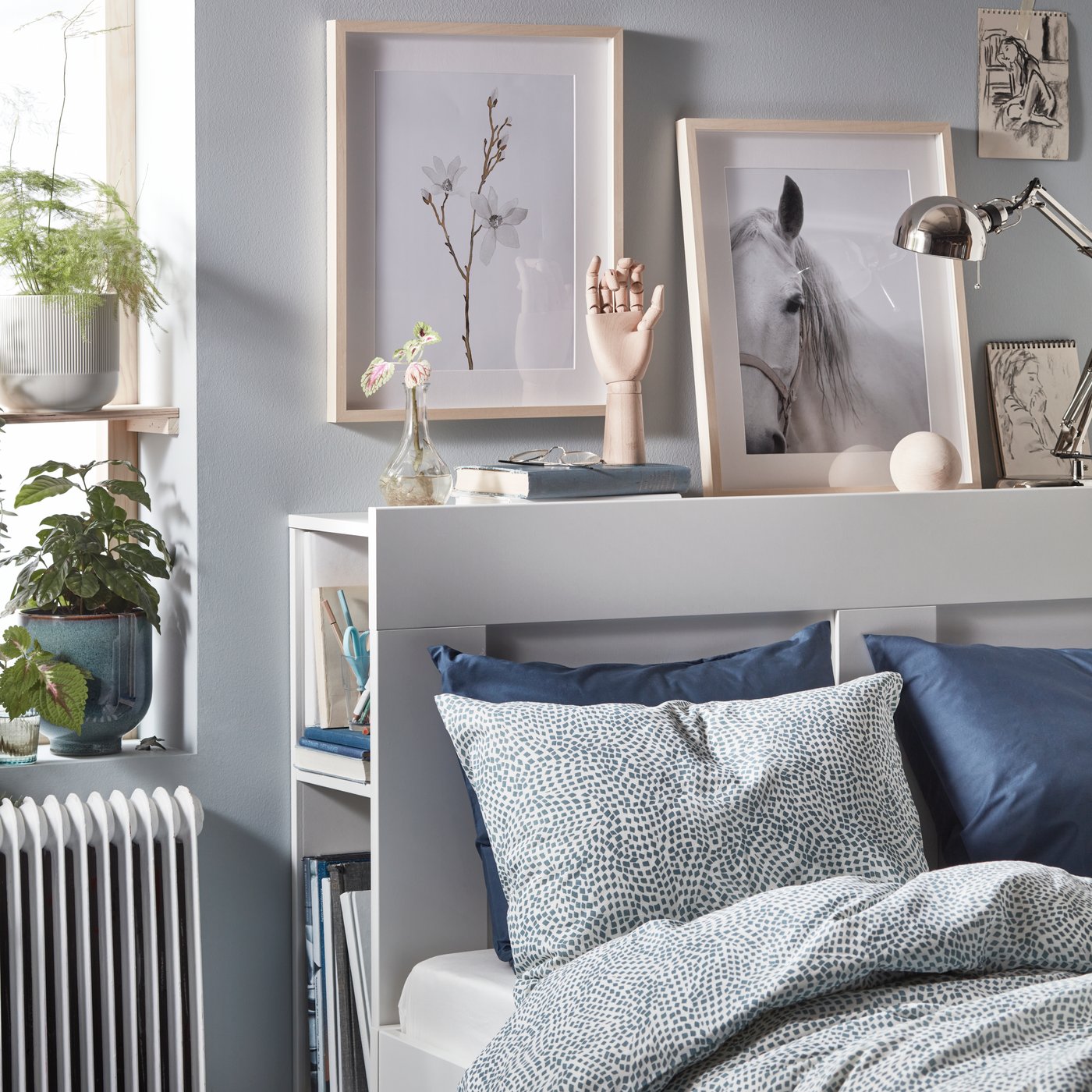 Cama BRIMNES blanca con un respaldo que tiene almacenaje incorporado. Además, tiene cuadros y decoración en la parte superior.
