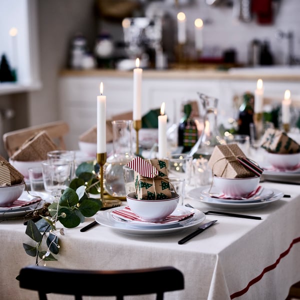 Une table de Noël chaleureuse est dressée avec des décorations VINTERFINT, des bougies allumées et de petits cadeaux emballés dans des coupes.