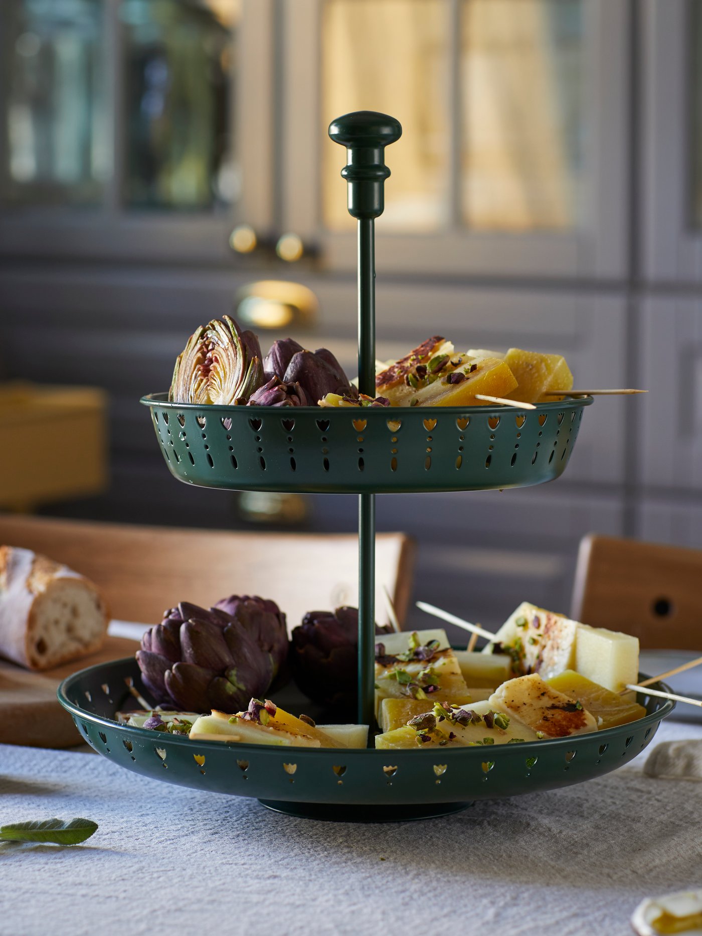 A GARNERA two-tier serving stand in dark green on a table, the serving stand holding various types of food such as cheeses.