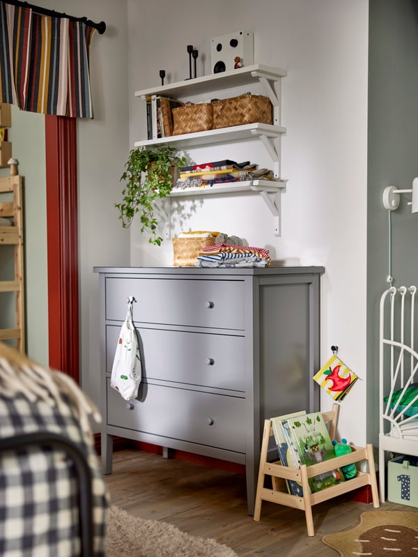 Grey chest of three drawers with three wall mounted shelfs above, baskets, plants and decoration around.