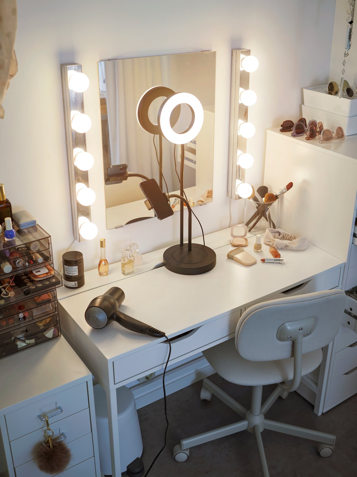 A brightly lit ALEX table in the bedroom with a mirror and two rows of lights next to it. A selfie handphone stand sits in the middle of the table, and other vanity items are placed on the table.