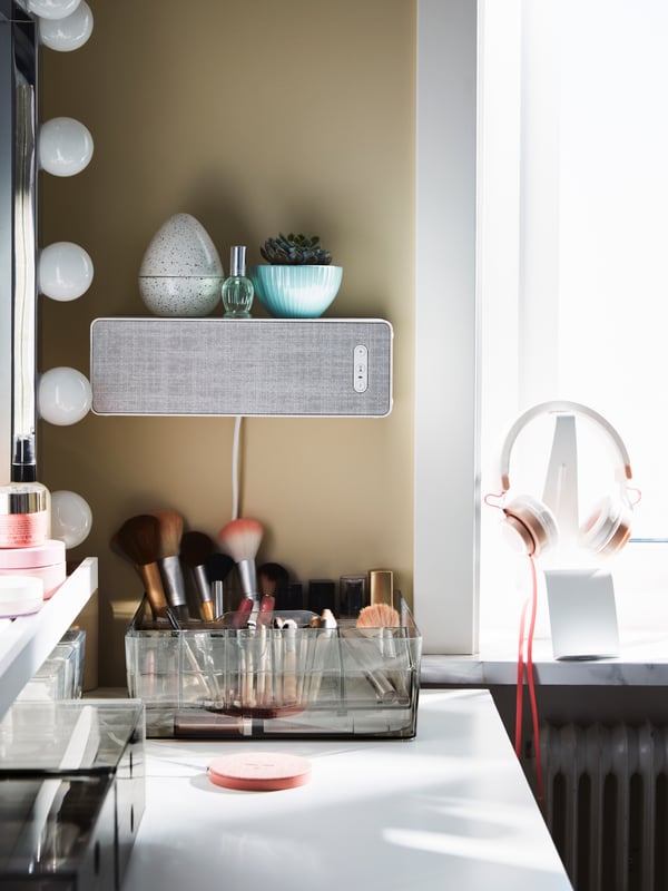 Une enceinte SYMFONISK blanche sur le mur au-dessus d’un bureau blanc avec des produits de maquillage et des pinceaux dans des organiseurs transparents.