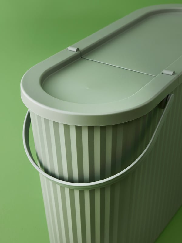 The ribbed side, rounded corner, lid and folding handle of the grey-green DAMMÄNG 48 l bin with lid in a green space.