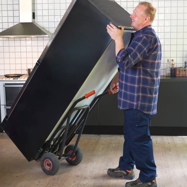 Une personne en tenue de travail utilise un chariot pour déplacer un réfrigérateur noir d'une cuisine carrelée.